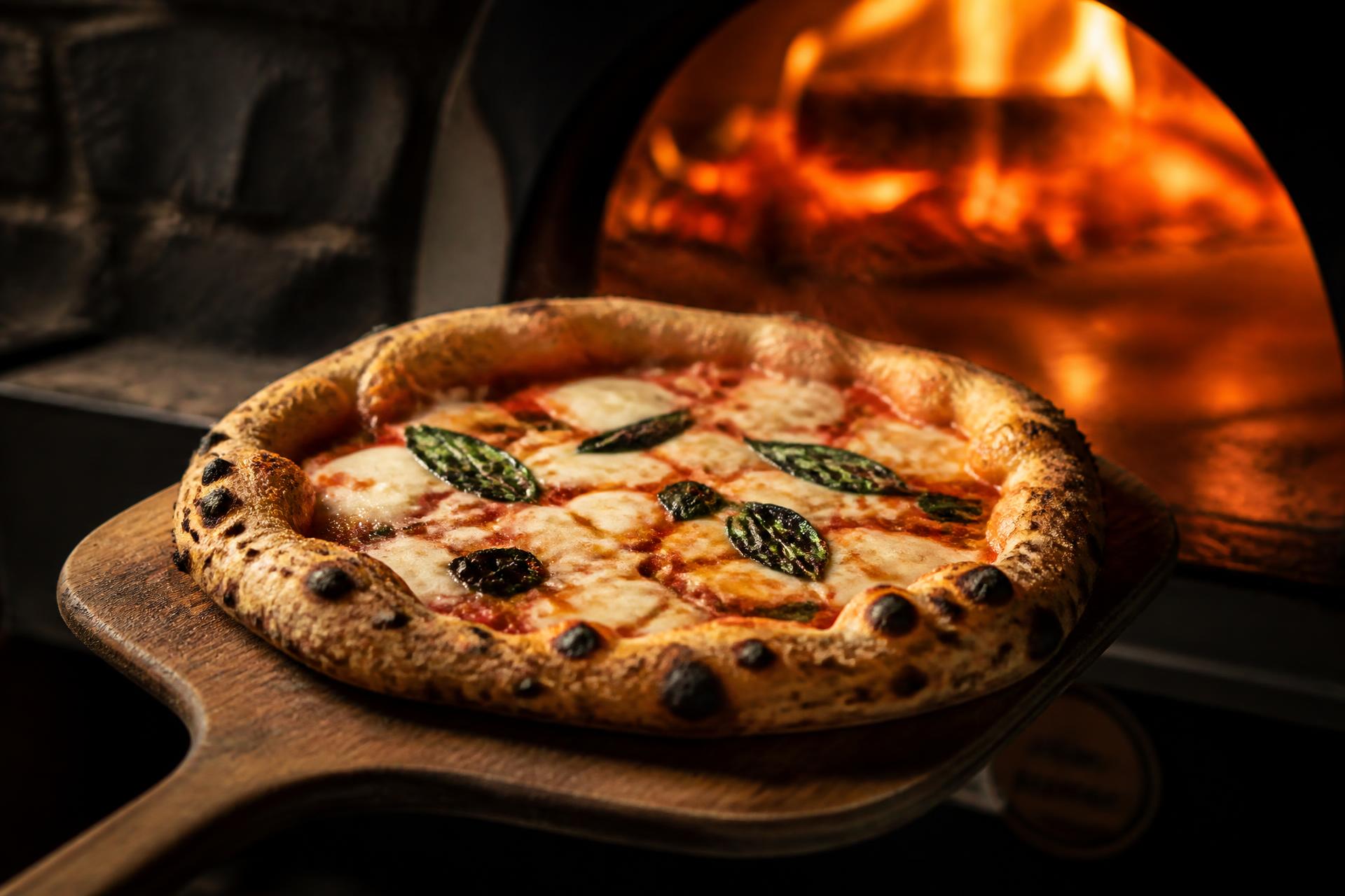 Wood-fired margherita pizza on a wooden peel in front of a blazing oven